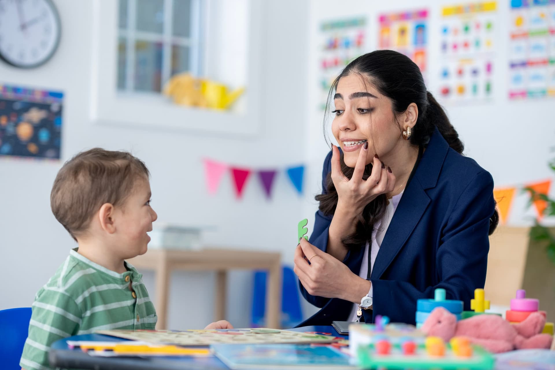 Latina BCBA clinician in professional attire modeling articulation with a green letter shape for a young toddler boy during an ABA therapy session in a bright educational room with colorful educational posters