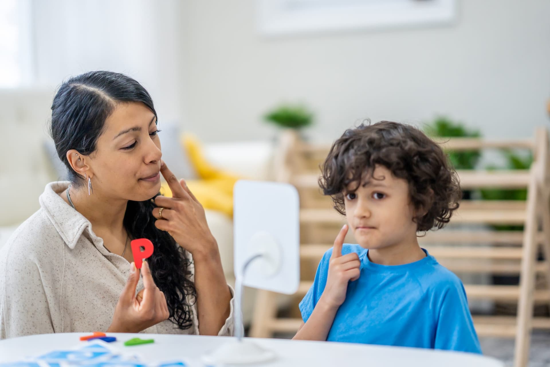 Latina behavior therapist holding a red letter P and touching her cheek to model articulation while a young Hispanic boy with curly hair mirrors the gesture using a mirror during a skill acquisition session at home
