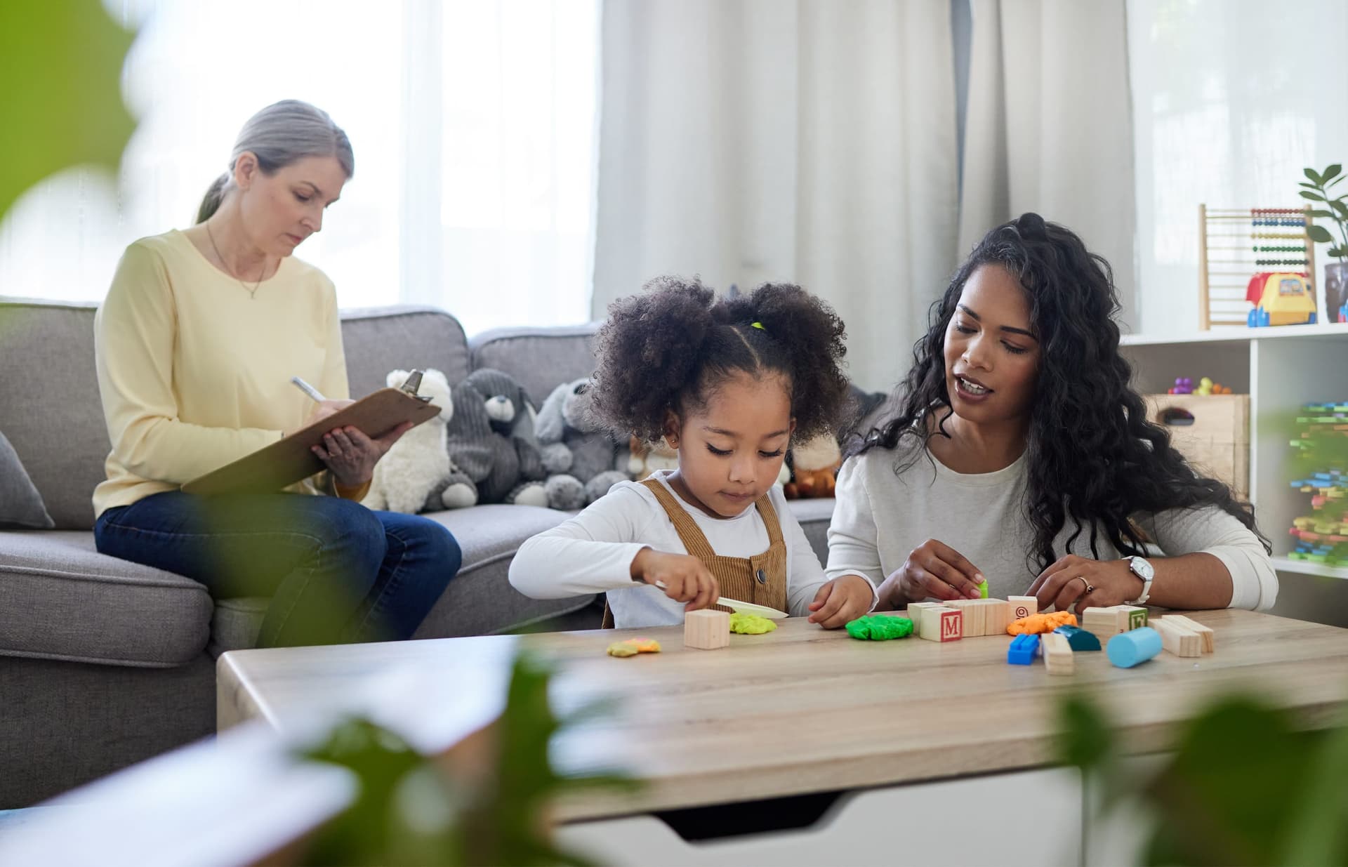 BCBA supervisor taking notes on a clipboard while observing a behavior technician conducting a naturalistic teaching session with a young mixed-race girl using letter blocks and play-doh on a coffee table in a home therapy setting