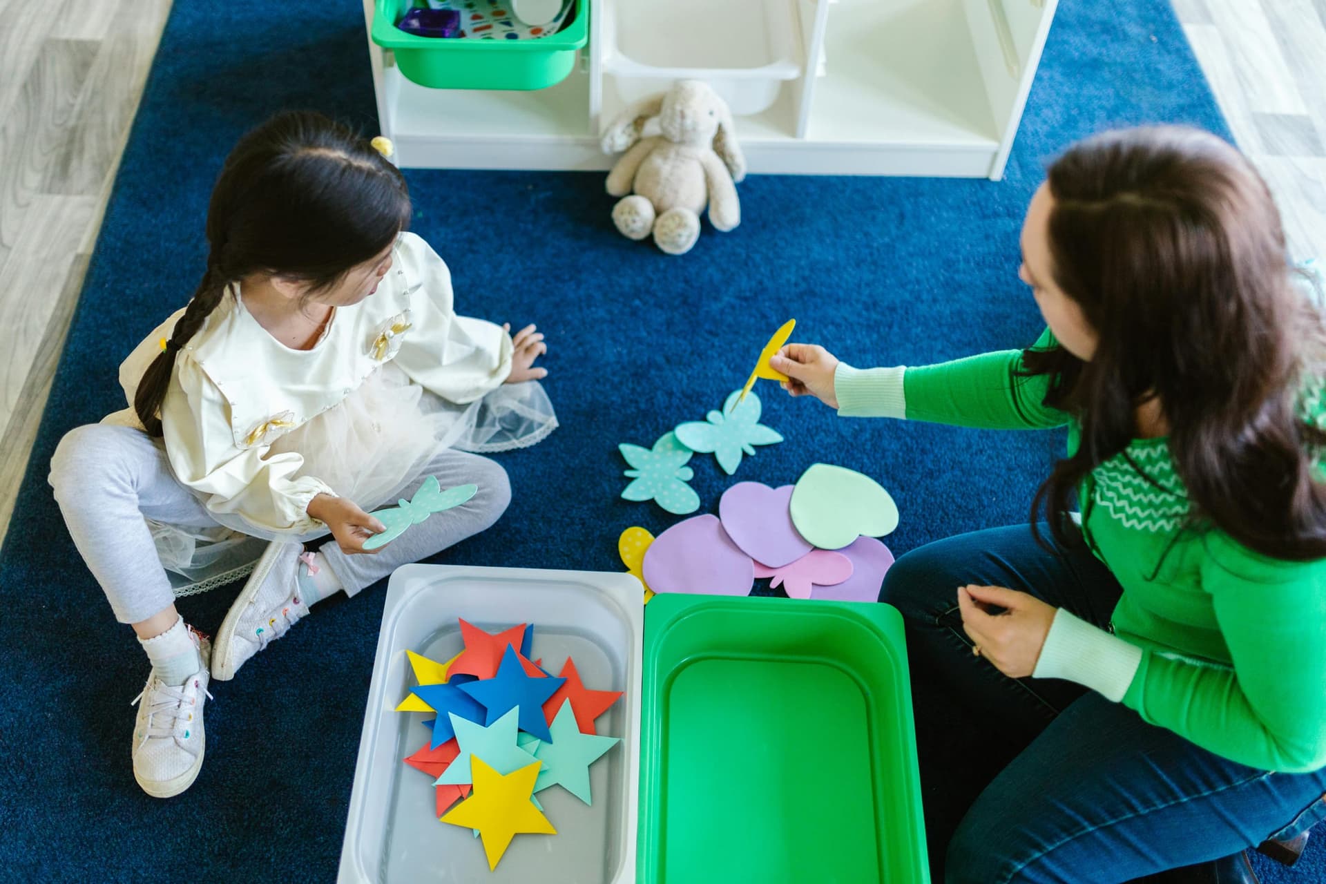 Behavior therapist and a young girl sitting on a blue carpet sorting colorful foam shapes during an ABA early intervention session